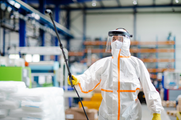 Front view portrait of man worker with protective mask and suit disinfecting industrial factory with spray gun.