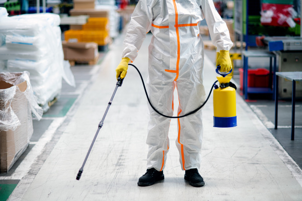 Front view portrait of man worker with protective mask and suit disinfecting industrial factory with spray gun.