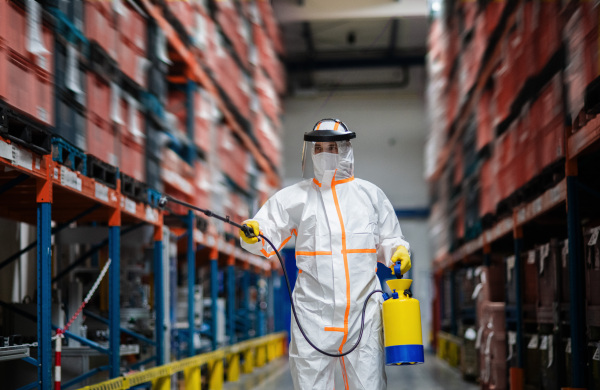 Front view portrait of man worker with protective mask and suit disinfecting industrial factory with spray gun.