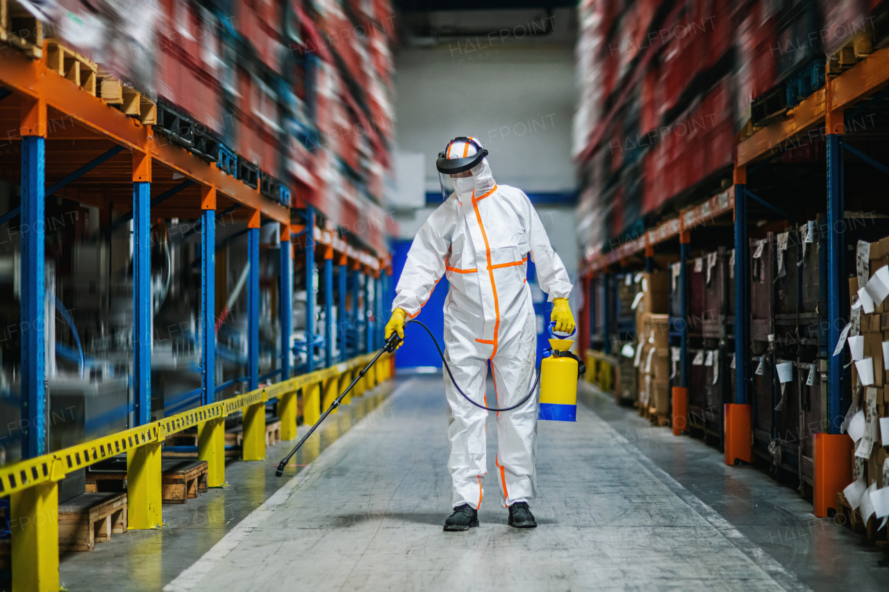 Front view portrait of man worker with protective mask and suit disinfecting industrial factory with spray gun.