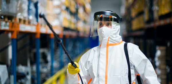 Front view portrait of man worker with protective mask and suit disinfecting industrial factory with spray gun.