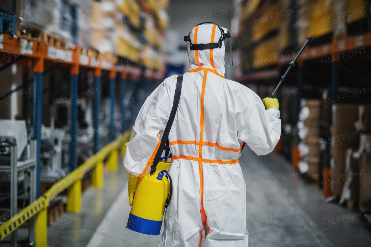 Rear view of man worker with protective mask and suit disinfecting industrial factory with spray gun.