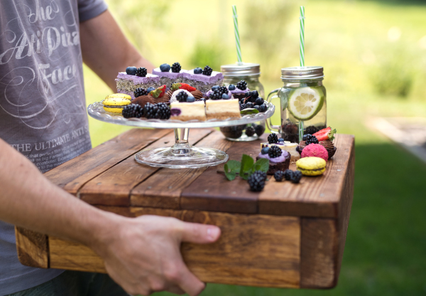 Unrecognizable man carrying outdoors selection of colorful and delicious cake desserts and drinks on tray.