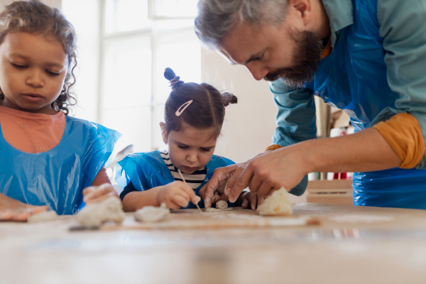 Little kids with a teacher working with pottery clay during creative art and craft class at school.