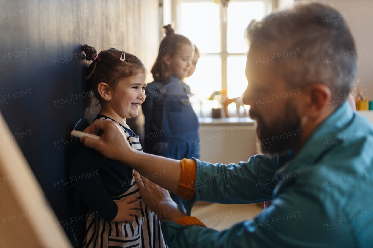 A teacher drawing little girl with chalks on blackboard wall indoors in playroom.