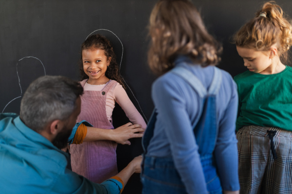 A teacher drawing little girl student with chalks on blackboard wall indoors in playroom.