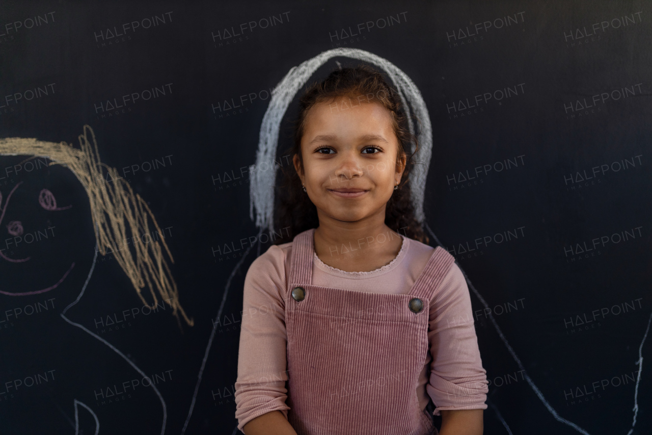 A little girl standing against blackboard with drawing indoors in playroom, looking at camera.