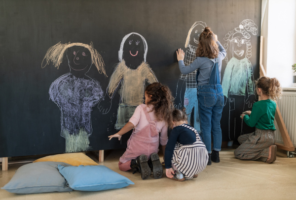 A rear view of group of little girls drawing with chalks on blackboard wall indoors in playroom.