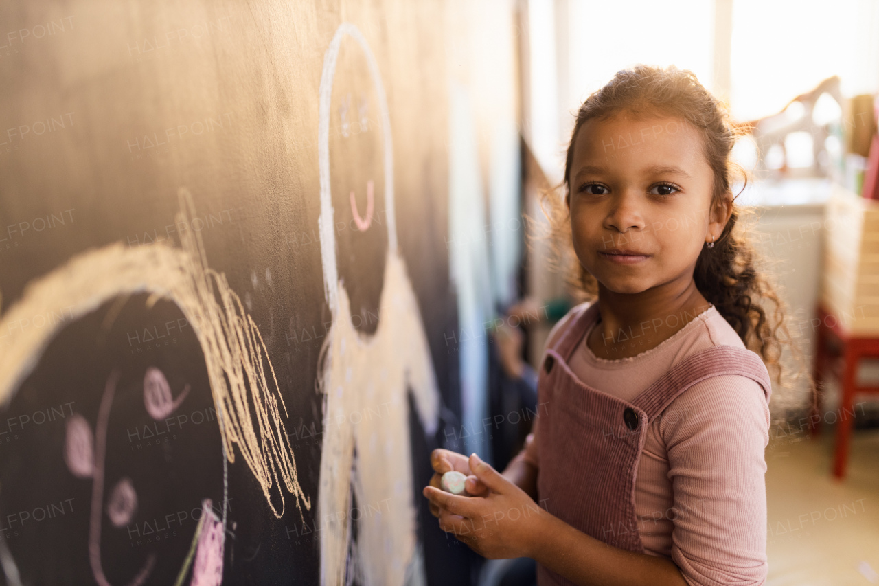 A little girl drawing with chalks on blackboard wall indoors in playroom, looking at camera.