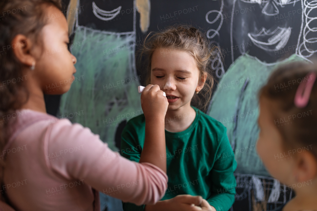 A little girl drawing with chalks on other girl's cheeks indoors in playroom.