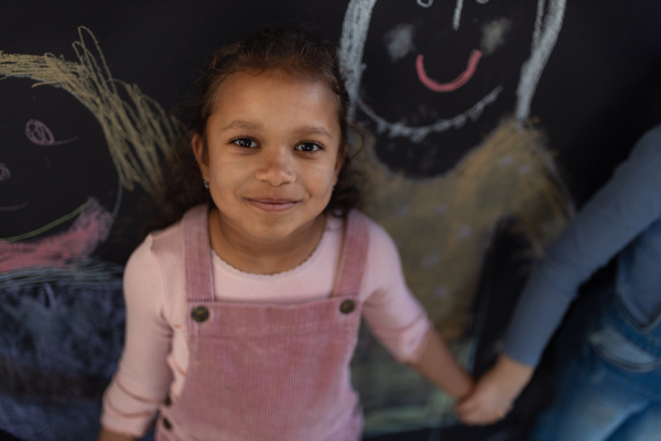 Little girl posing and smiling in front of blackboard wall drawings indoors in playroom.