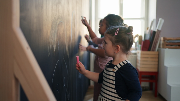 A little girl drawing her friends with chalks on blackboard wall indoors in playroom.