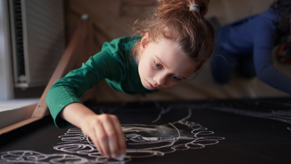 Little girls drawing with chalks on a blackboard wall indoors in playroom.