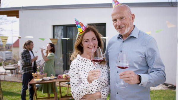 A mature couple with wine outdoors in garden at home, looking at camera. Birthday celebration party.