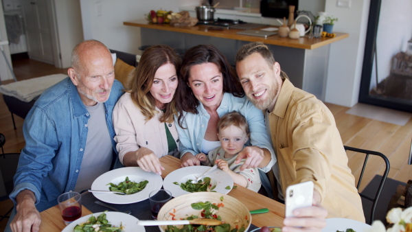 Happy multigeneration family indoors at home eating healthy lunch, taking a video with smartphone.