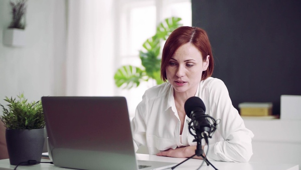 A front view of young woman indoors, recording a podcast at home. Slow motion.