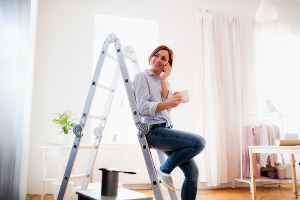 A portrait of young creative woman with a cup of coffee and smartphone painting wall black. A startup of small business.