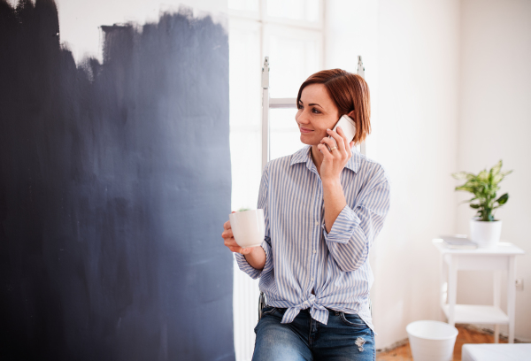 A portrait of young creative woman with a cup of coffee and smartphone painting wall black. A startup of small business.