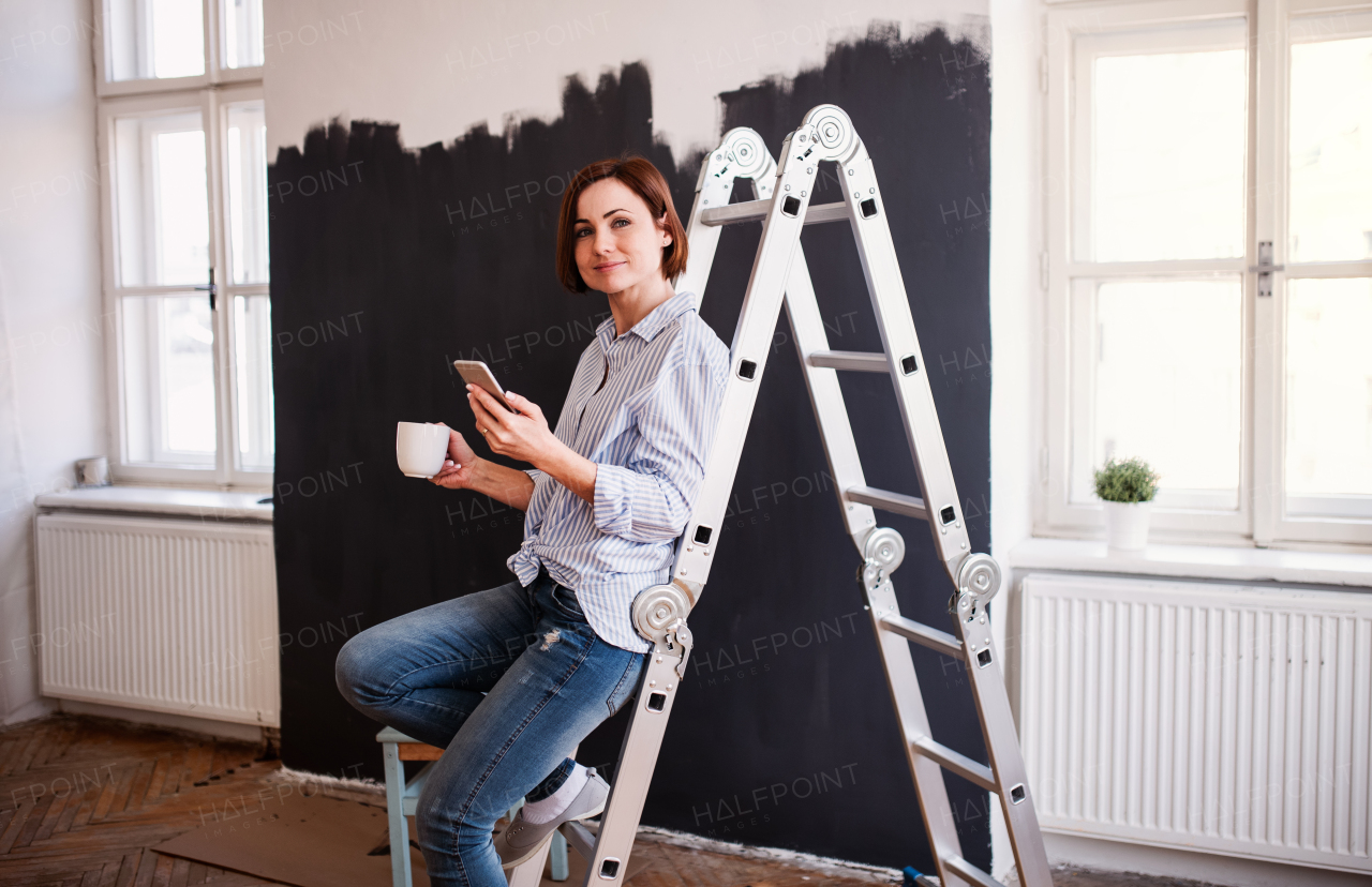 A portrait of young creative woman with a cup of coffee and smartphone painting wall black. A startup of small business.