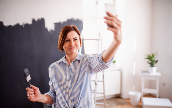 A portrait of young creative woman with smartphone painting wall black, taking selfie. A startup of small business.