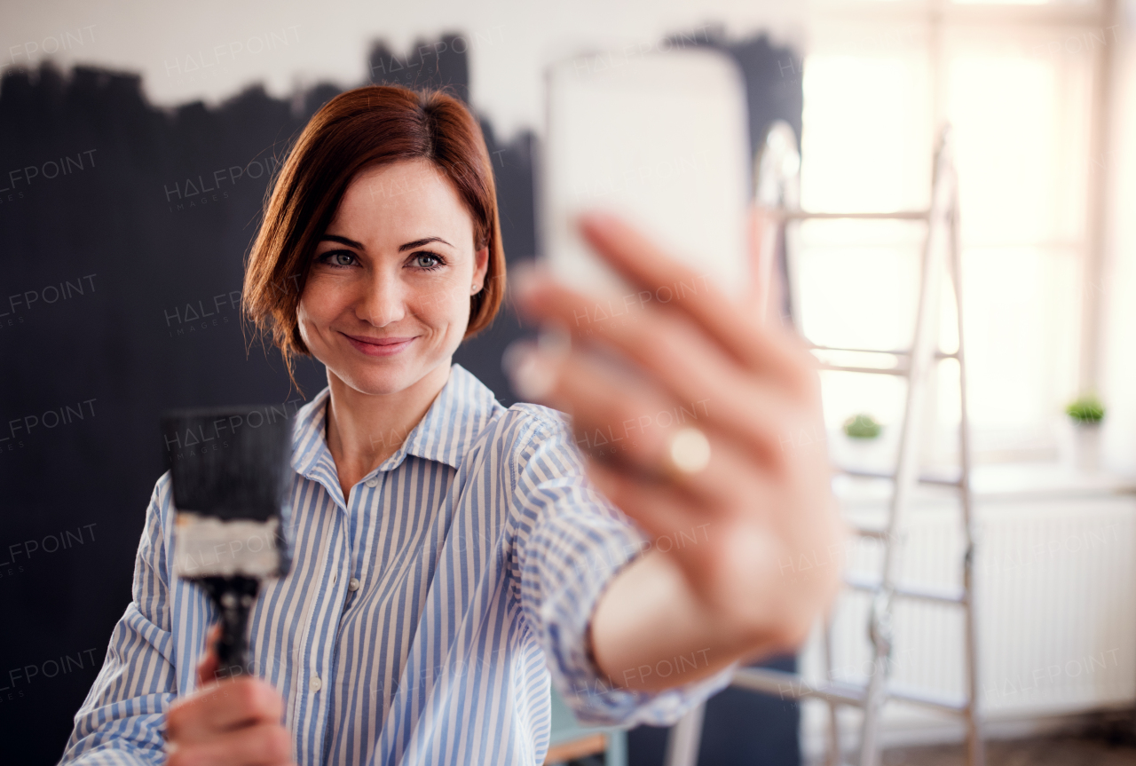 A portrait of young creative woman with smartphone painting wall black, taking selfie. A startup of small business.