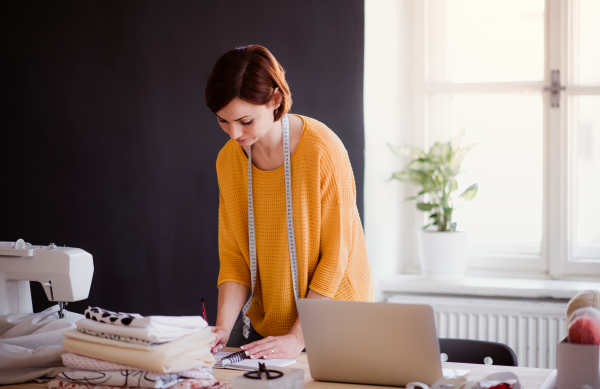 Young creative woman with laptop working in a studio, startup of small tailoring business.