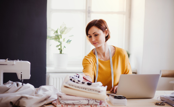 Young creative woman with laptop working in a studio, startup of small tailoring business.