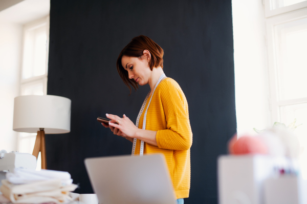 Young creative woman in a studio, using smartphone. A startup of small tailoring business. Copy space.