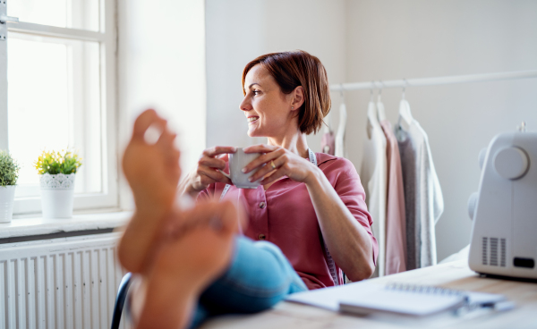 Young creative woman with coffee in a studio, resting with feet on desk. A startup of small tailoring business.