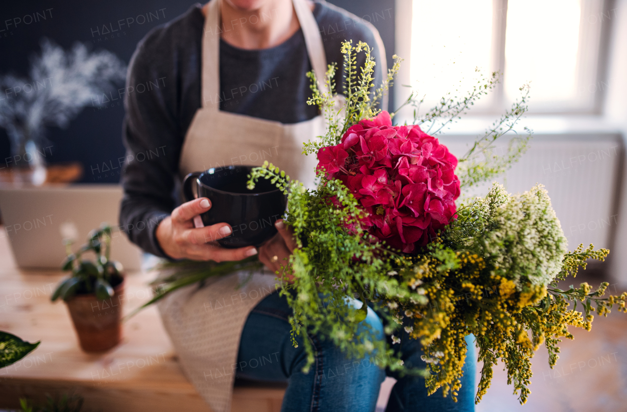 A midsection of young creative woman with a coffee arranging flowers in a flower shop. A startup of florist business.