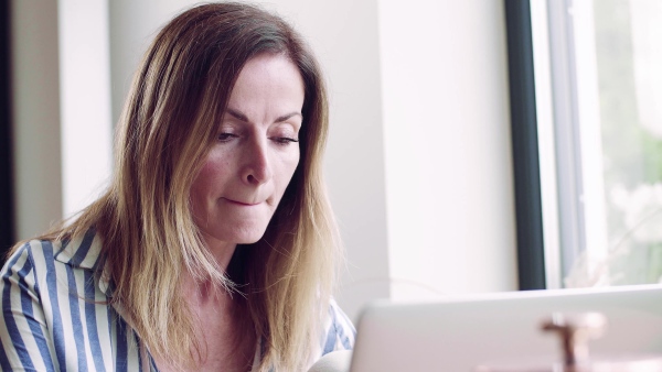 A confident mature woman sitting at the table, working with laptop in home office.