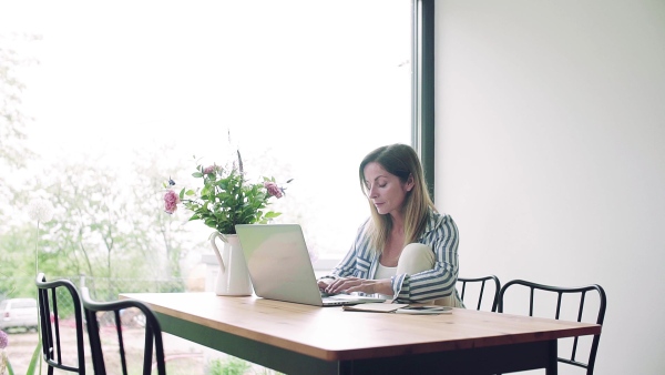 A mature woman with coffee and laptop indoors working in home office. Slow motion.