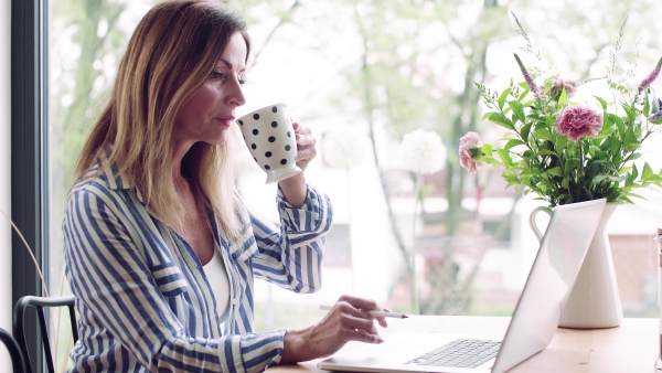 A confident mature woman sitting at the table, working with laptop in home office.