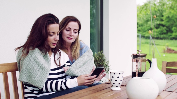 A mother with adult daughter sitting at the table outdoors on a terrace, using smartphone.