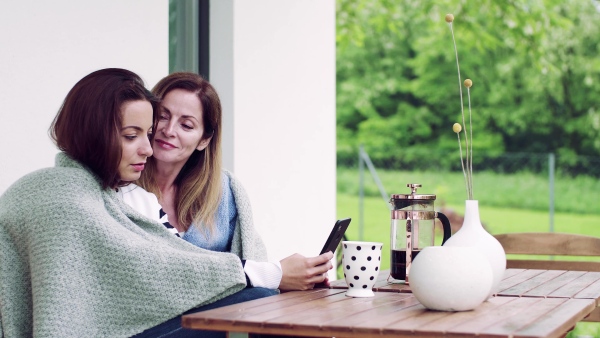 A mother with adult daughter sitting at the table outdoors on a terrace, using smartphone.