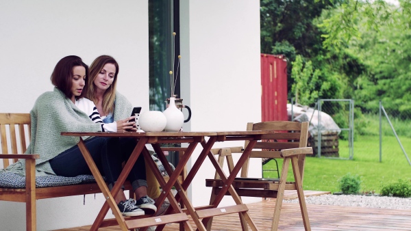 A mother with adult daughter sitting at the table outdoors on a terrace, using smartphone.