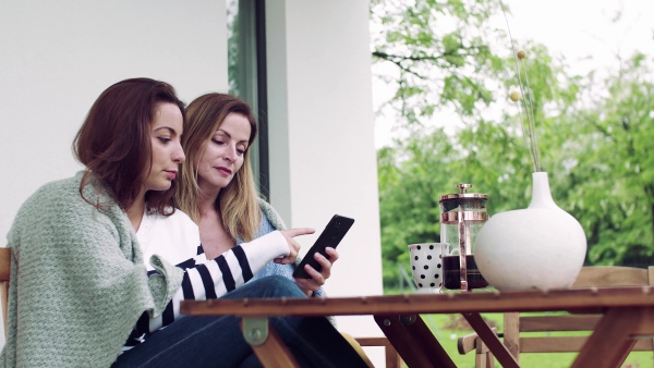 A mother with adult daughter sitting at the table outdoors on a terrace, using smartphone.