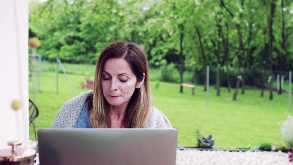 A front view of mature woman sitting at the table on a terrace outdoors, working with laptop.