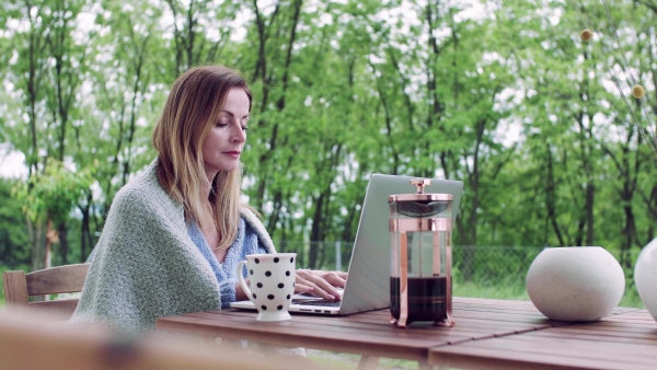 A mature woman sitting at the table on a terrace outdoors, working with laptop.