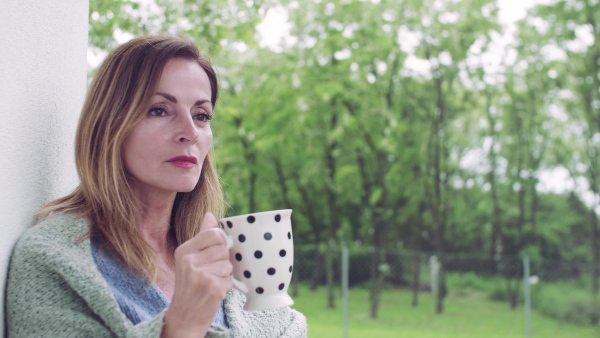 A mature woman with tea and blanket standing outdoors on terrace.