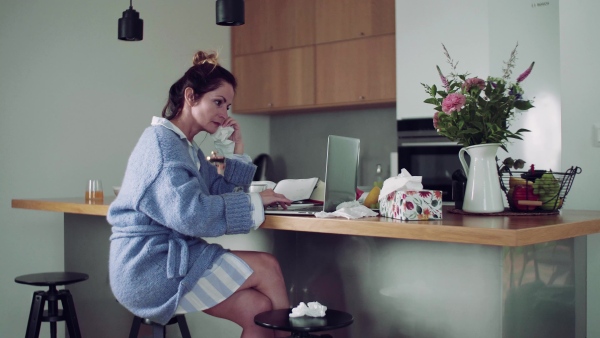 A sick mature woman with laptop sitting indoors, drinking tea when working in home office.