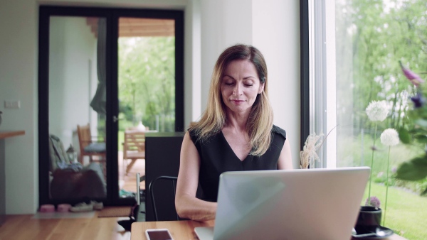 A confident mature woman sitting at the table, working with laptop in home office.