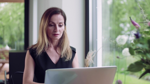 A confident mature woman sitting at the table, working with laptop in home office.