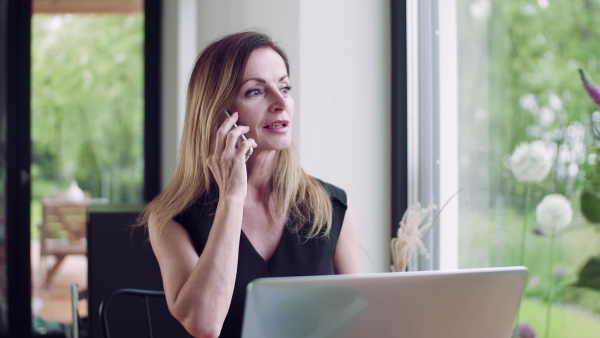 A mature woman with laptop sitting at the table, using smartphone in home office.