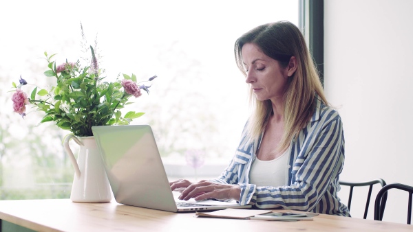 A confident mature woman sitting at the table, working with laptop in home office.