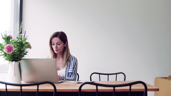 A confident mature woman sitting at the table, working with laptop in home office.