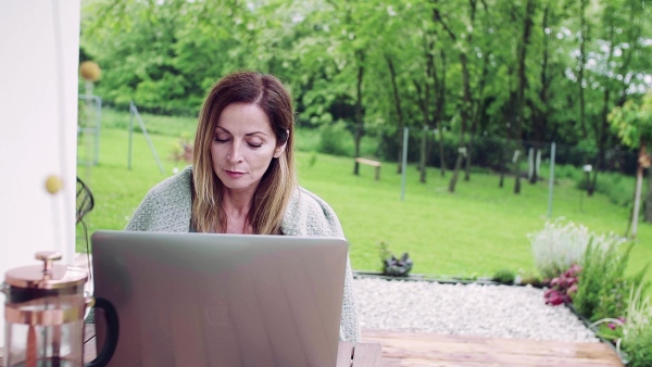 A front view of mature woman sitting at the table on a terrace outdoors, working with laptop. Slow motion.