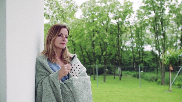 A mature woman with tea and blanket standing outdoors on terrace. Slow motion.