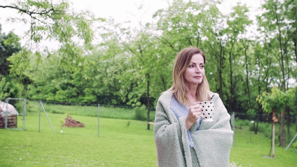 A sick mature woman with tea and blanket walking outdoors in the garden. Slow motion.