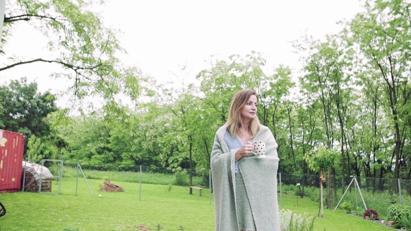 A sick mature woman with tea and blanket walking outdoors in the garden. Slow motion.